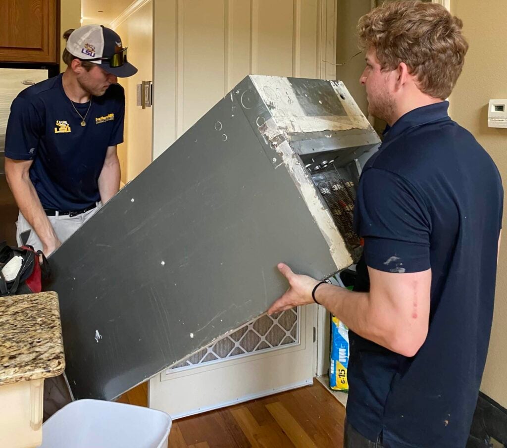 Two technicians in navy uniforms installing a large HVAC unit in a residential kitchen with granite countertops.