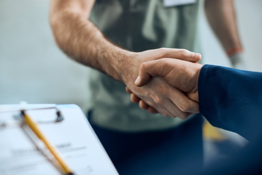 Two men shaking hands during a business meeting with a clipboard and pen visible, sealing a service agreement.